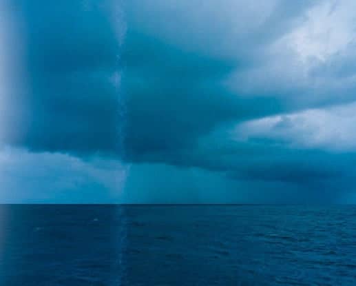 A vertical column of rain descends from a dark, stormy sky above the ocean.